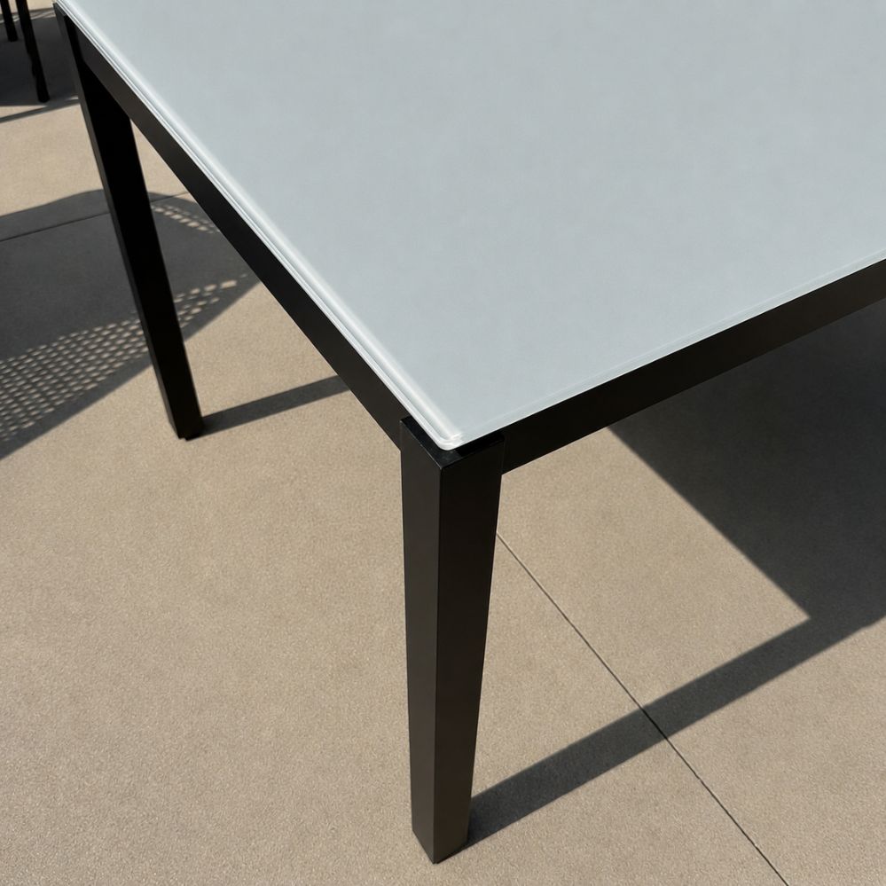 Detail van het lichtgrijze glazen tafelblad van 10 mm en het zwart mat aluminium frame van de Jati Kebon Juma tuintafel als terrasmodel in Temse