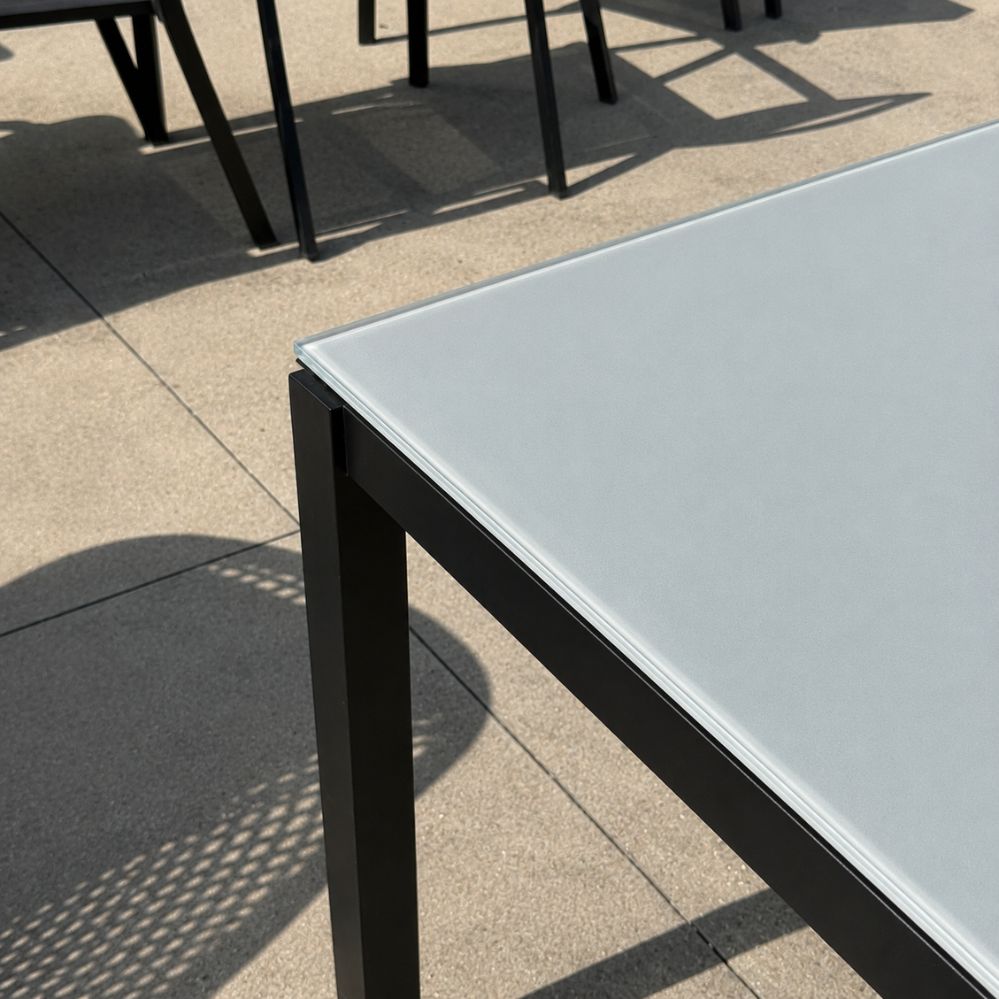 Detail van de hoek van het lichtgrijze glazen tafelblad en het zwart mat aluminium onderstel van de Jati Kebon Juma tuintafel als terrasmodel in Temse